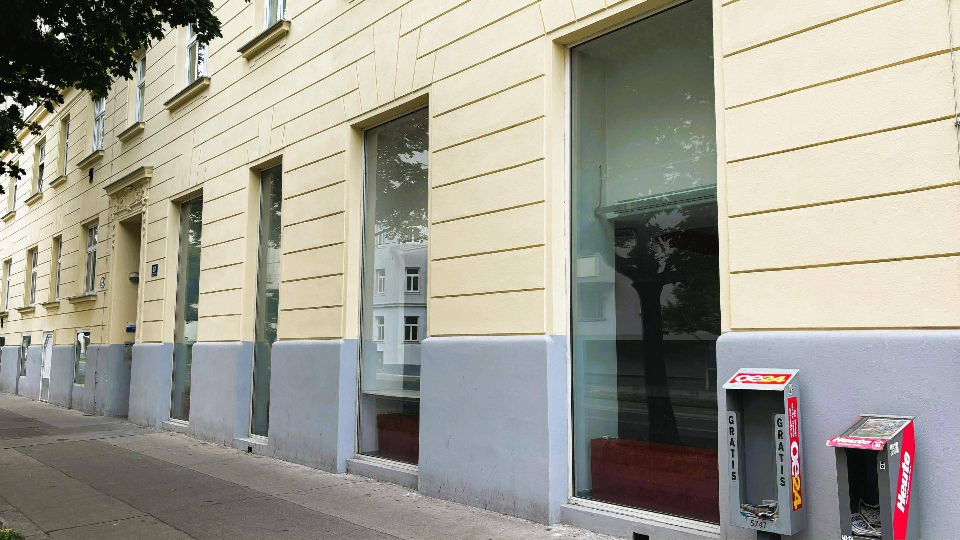 Nahaufnahme der Gebäudefassade im Erdgeschoss mit großen Fenstern und einem Zeitungsständer.