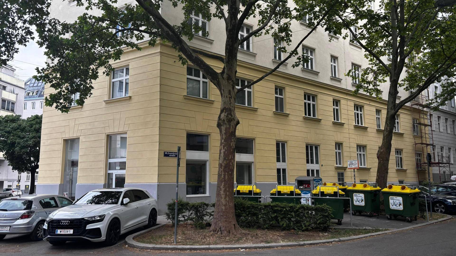 Eckgebäude mit parkenden Autos und Mülltonnen auf der Straße, umgeben von Bäumen.
