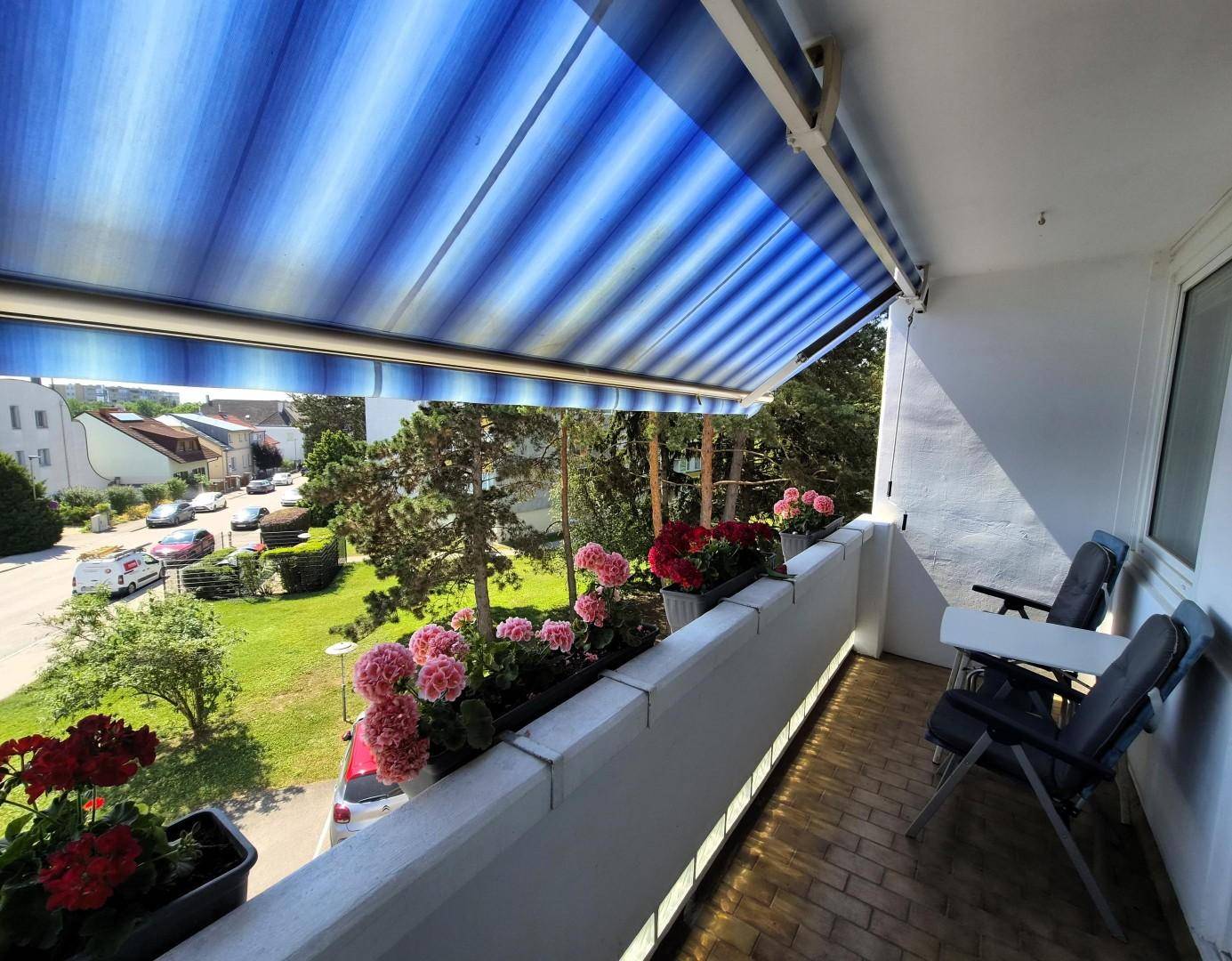 Gemütlicher Balkon mit blauer Markise, bunten Blumenkästen und Sitzgelegenheiten im Freien.