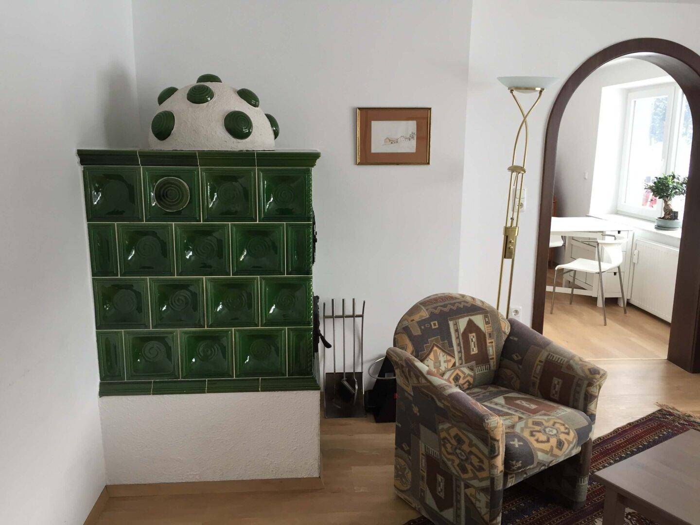 Gemütliches Wohnzimmer mit grünem Kachelofen und traditionellem Sessel auf Teppichboden.