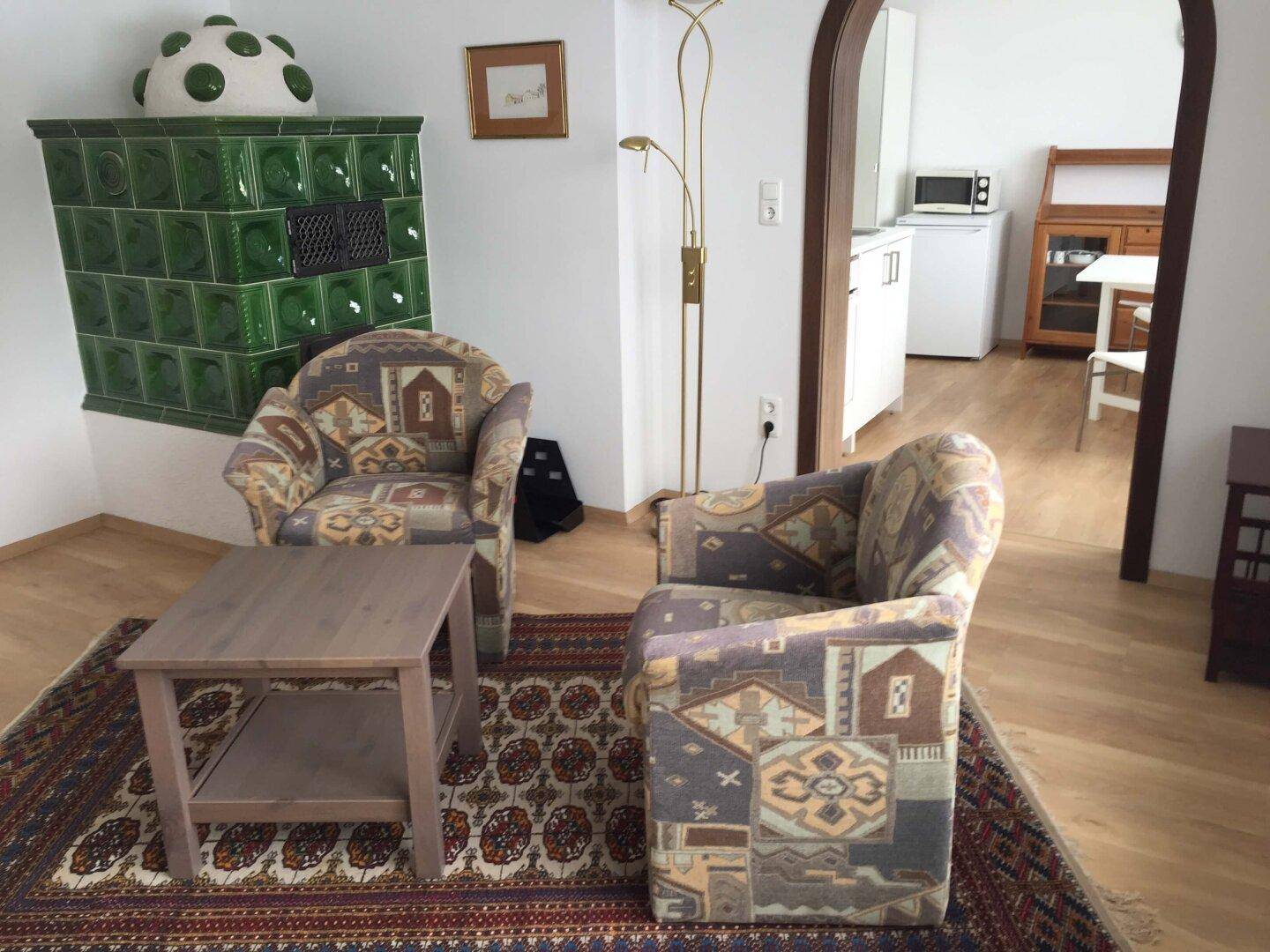 Wohnzimmer mit zwei gemusterten Sesseln, Couchtisch und grünem Kachelofen.