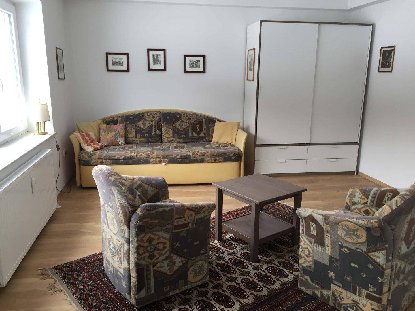 Helles Wohnzimmer mit Schlafsofa, Kleiderschrank und Holzboden.