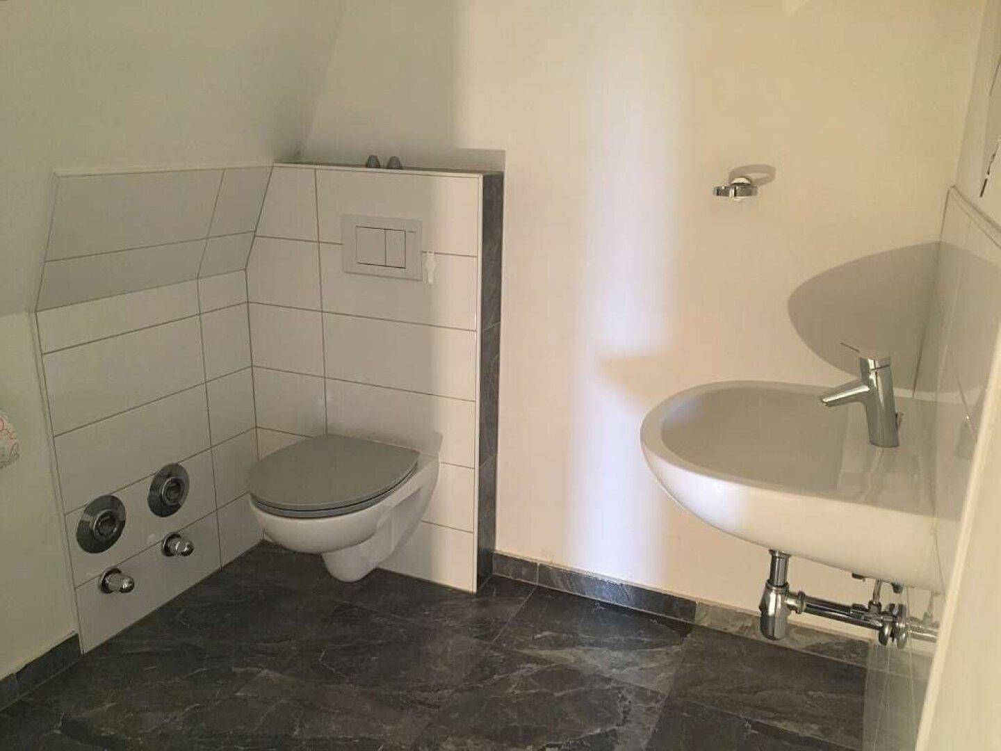 Modernes Gäste-WC mit Wand-WC und kleinem Waschbecken, gefliest mit dunklem Boden.
