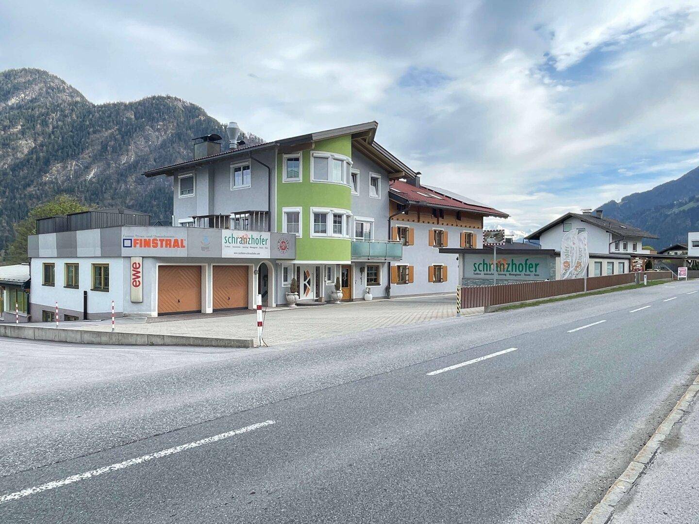 Mehrere Gewerbegebäude entlang einer Straße mit Bergen im Hintergrund unter bewölktem Himmel.