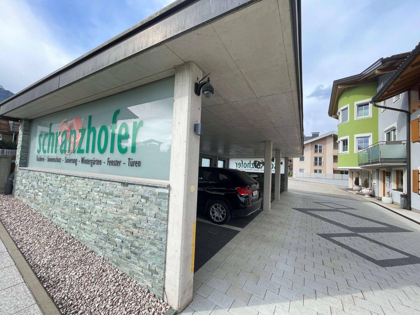 Überdachter Parkplatz mit Steinverkleidung und Werbeschild, daneben eine Straße und weitere Gebäude.