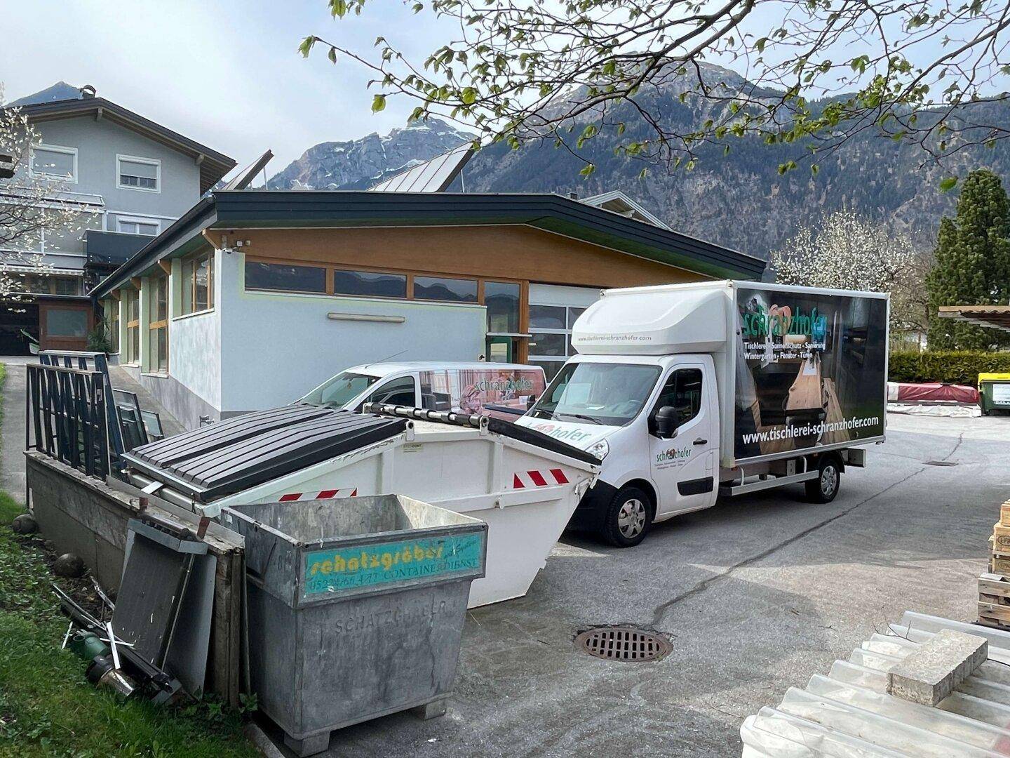 Außenbereich eines Gewerbegebäudes mit Lieferwagen, Müllcontainern und Bergen im Hintergrund.