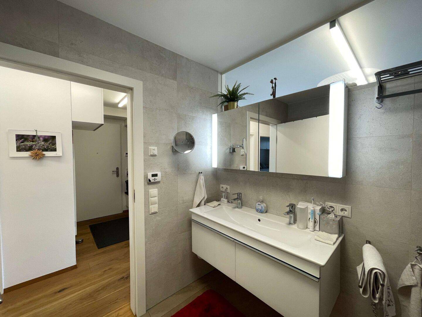 Modernes Badezimmer mit Waschtisch, beleuchtetem Spiegelschrank und grauen Fliesen, bietet direkten Zugang zum Flur.