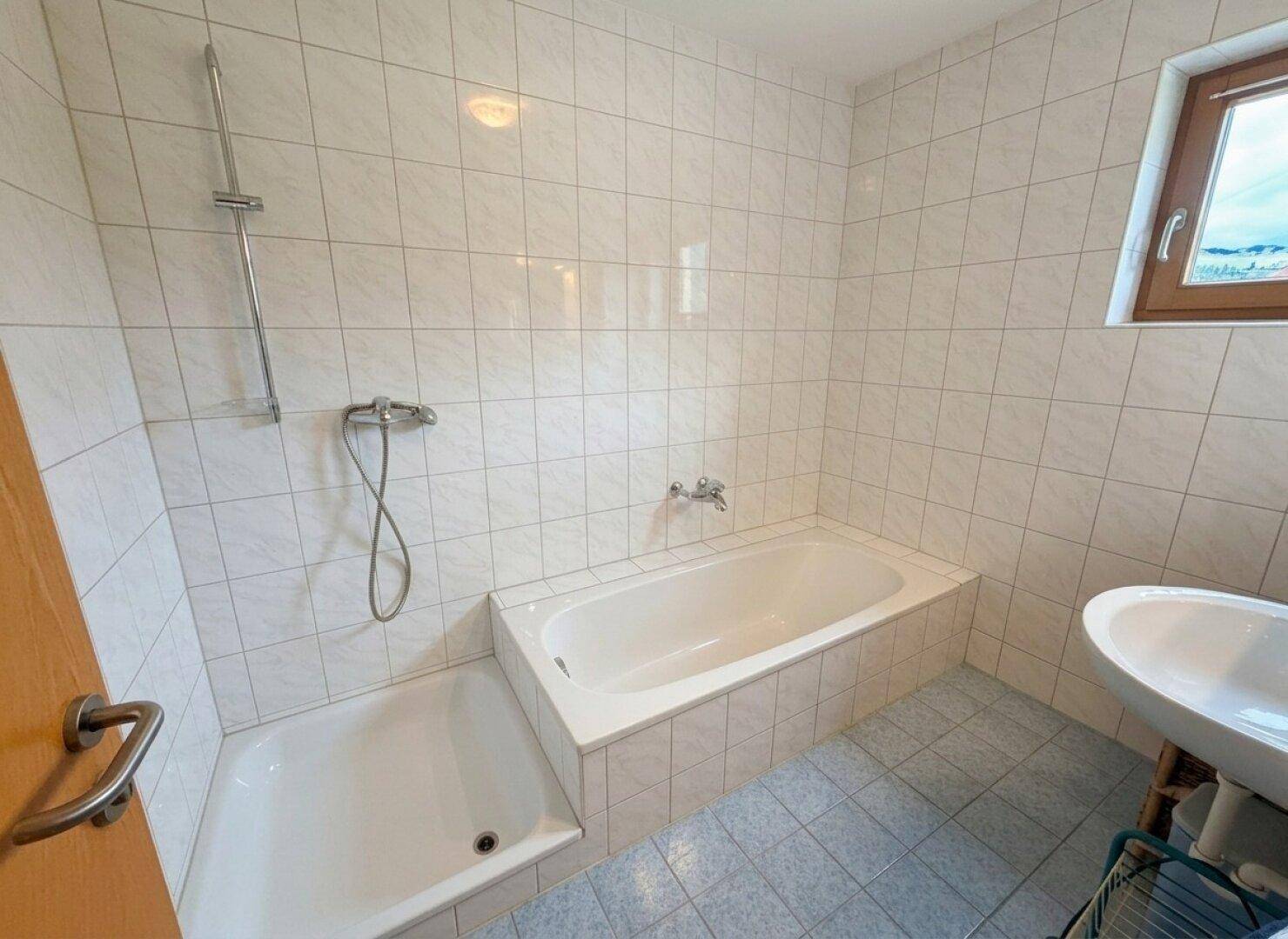 Gefliestes Badezimmer mit Badewanne, Dusche und Fenster mit Bergblick.