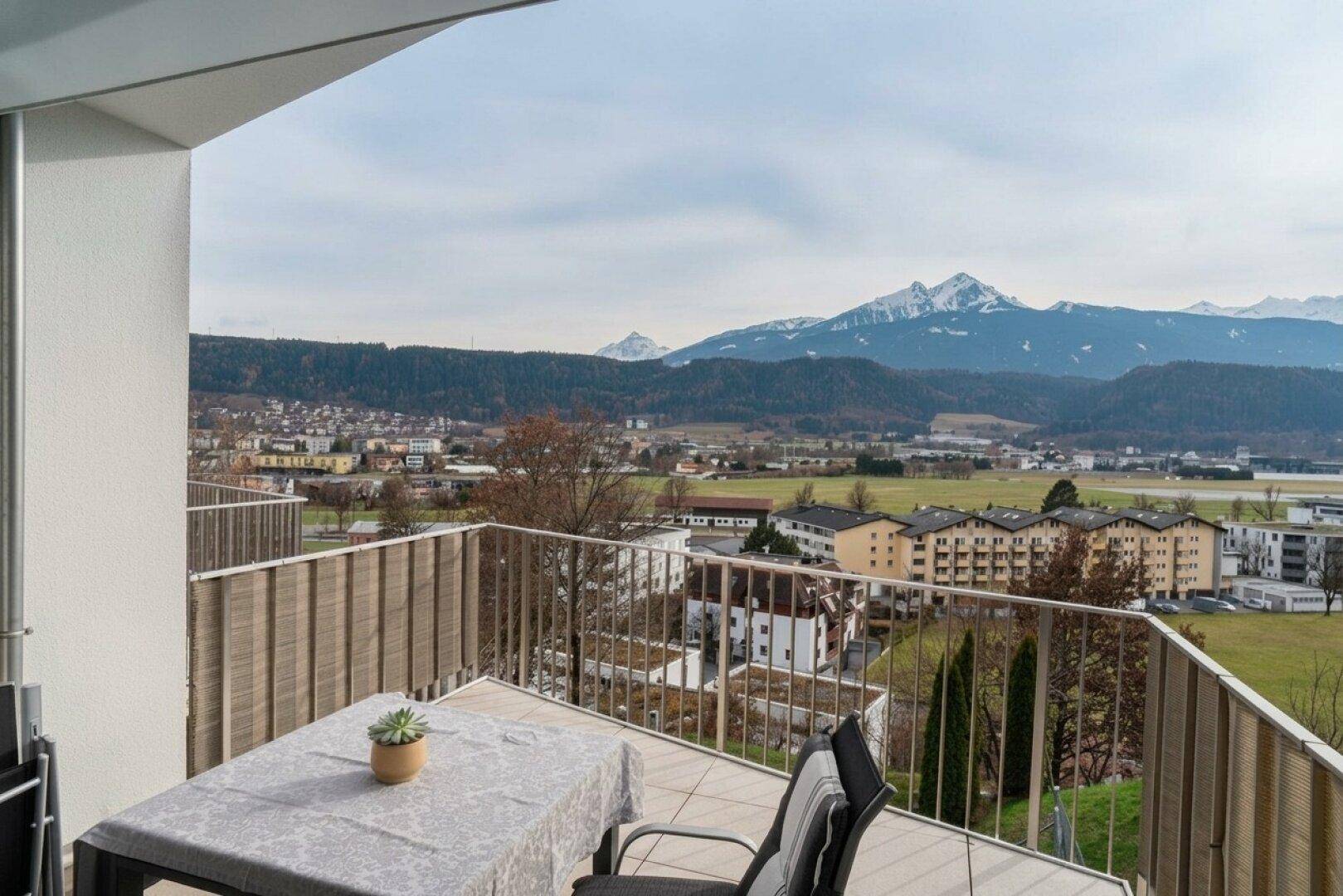 Möblierter Balkon mit Tisch und Stühlen, bietet einen Panoramablick auf die Stadt und die Alpen.
