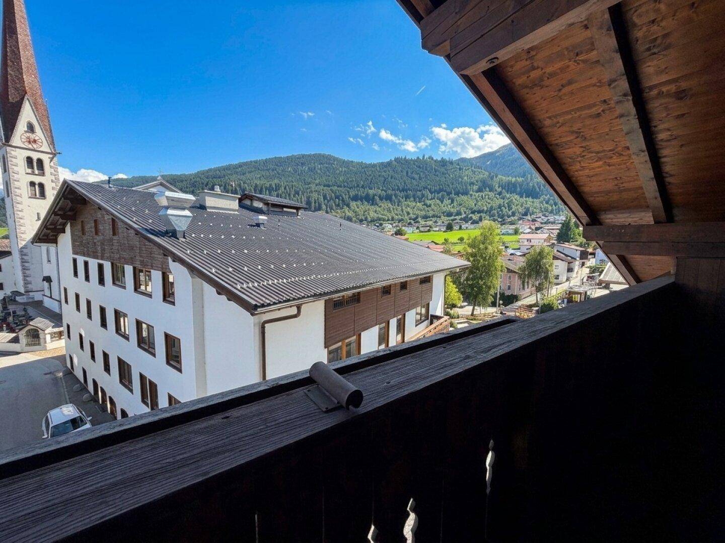 Balkon mit Holzelementen und weitem Blick über das Dorf und die umliegende Berglandschaft.