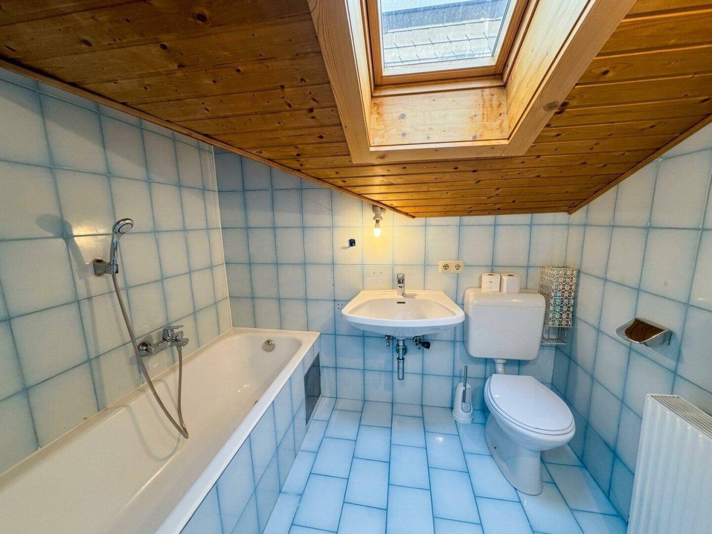 Badezimmer mit blauen Fliesen, Holzdecke, Badewanne, Waschbecken, Toilette und Dachfenster.