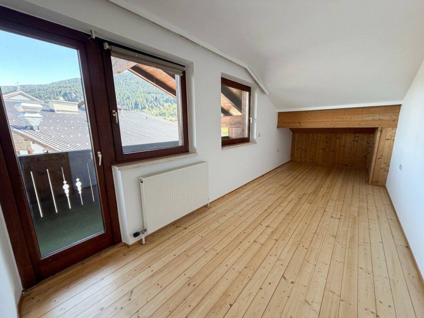 Geräumiger Raum mit Holzplankenboden, weißen Wänden und Zugang zu einem Balkon.