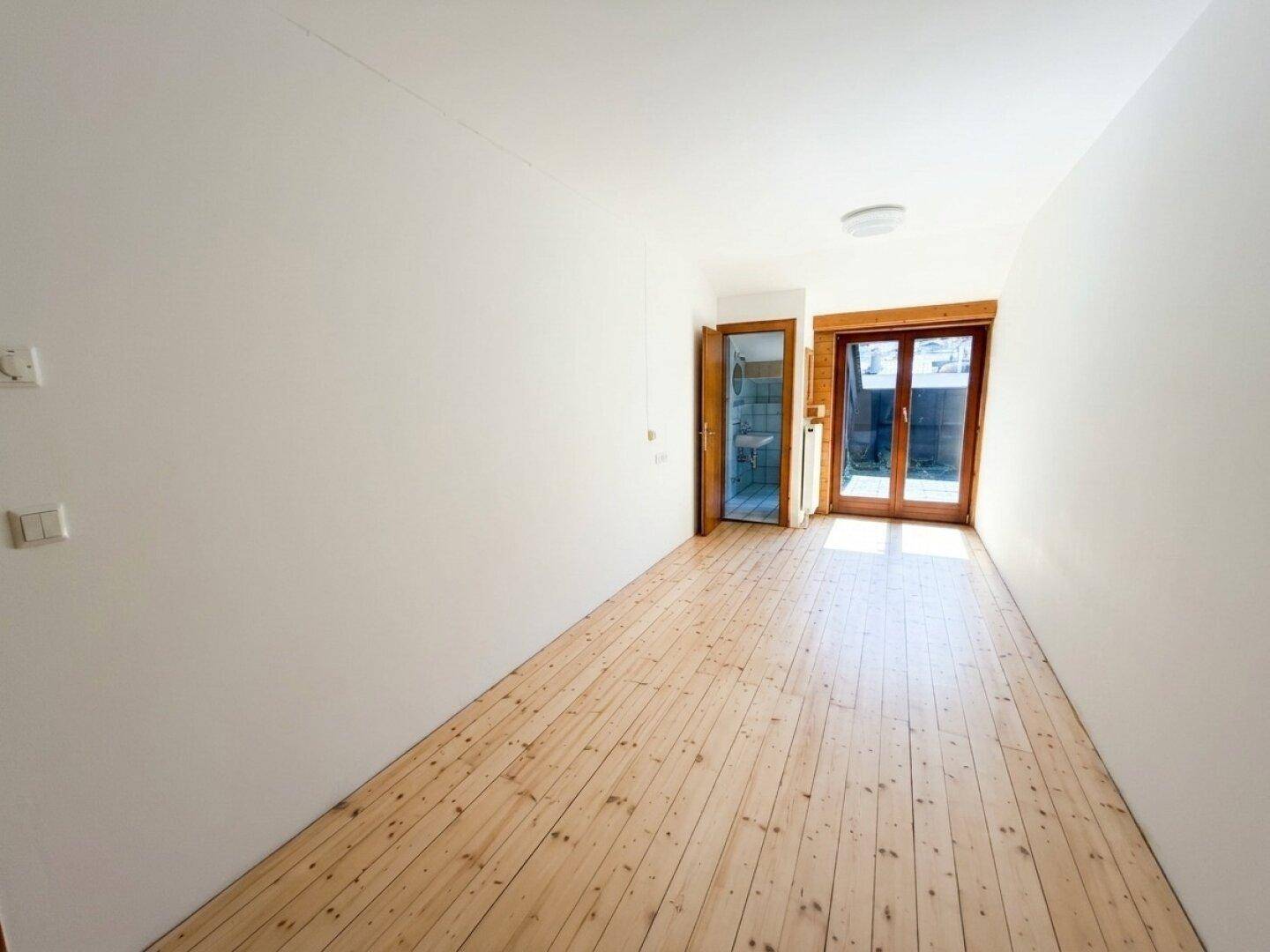 Heller, leerer Raum mit weißen Wänden, Holzdielenboden und Zugang zu einem Außenbereich.