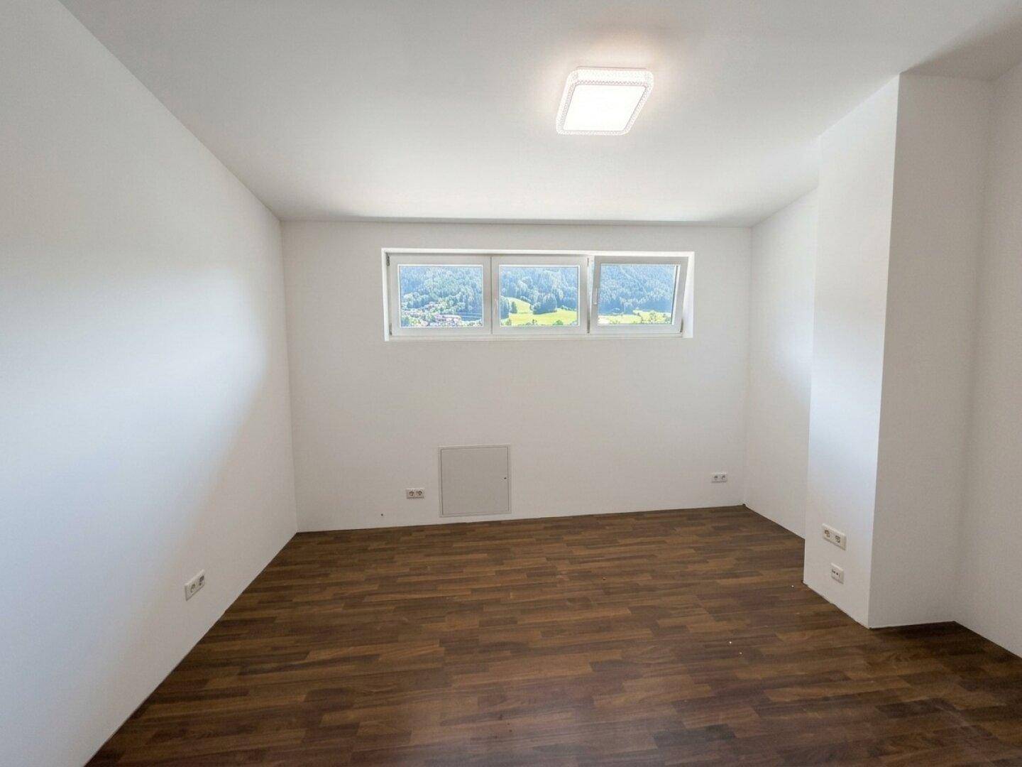 Leerer Raum mit weißen Wänden, dunklem Laminatboden und Blick auf grüne Hügel.