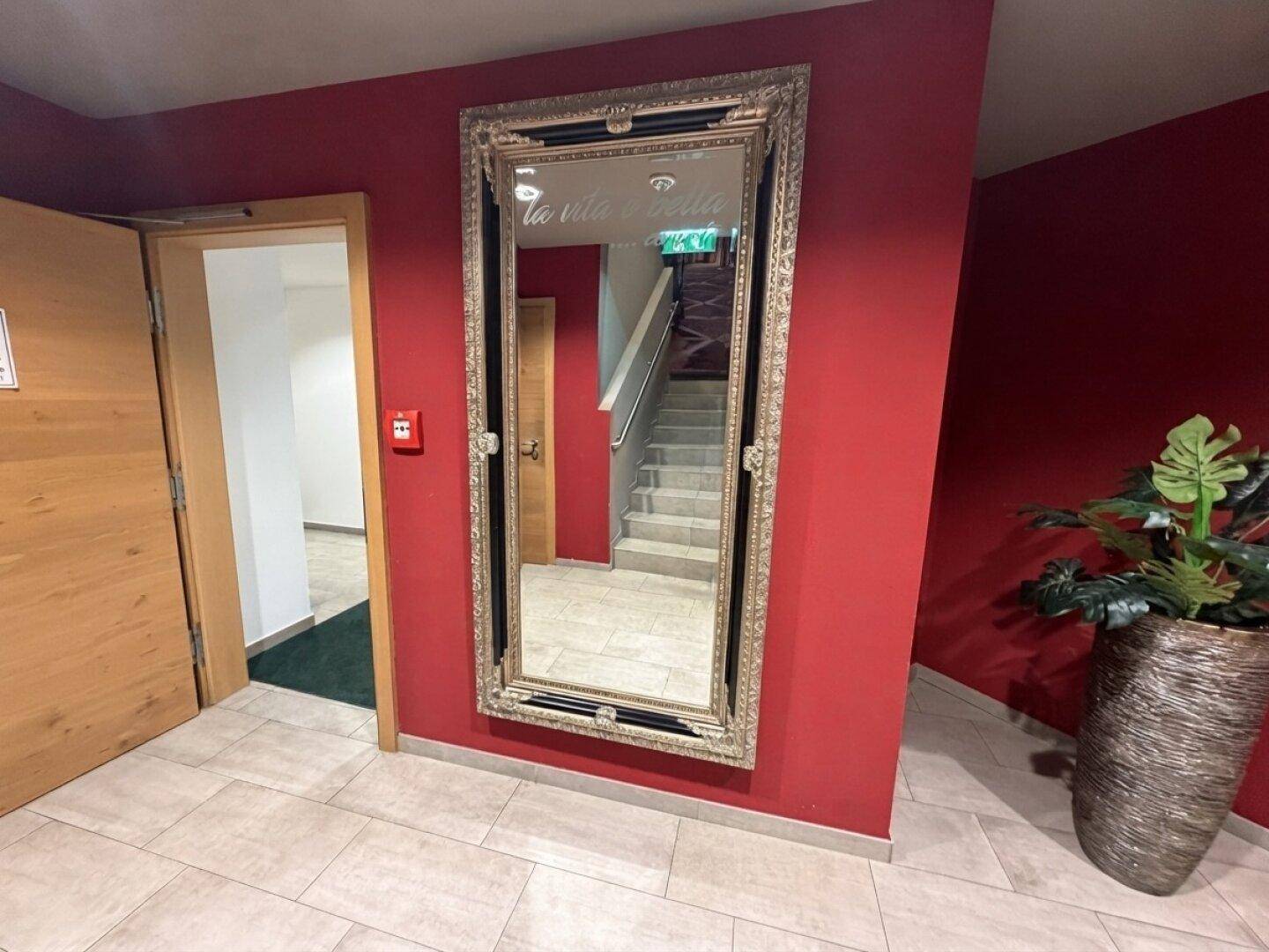 Flur mit roter Wand, einem großen Spiegel und Blick auf ein Treppenhaus im Hintergrund.