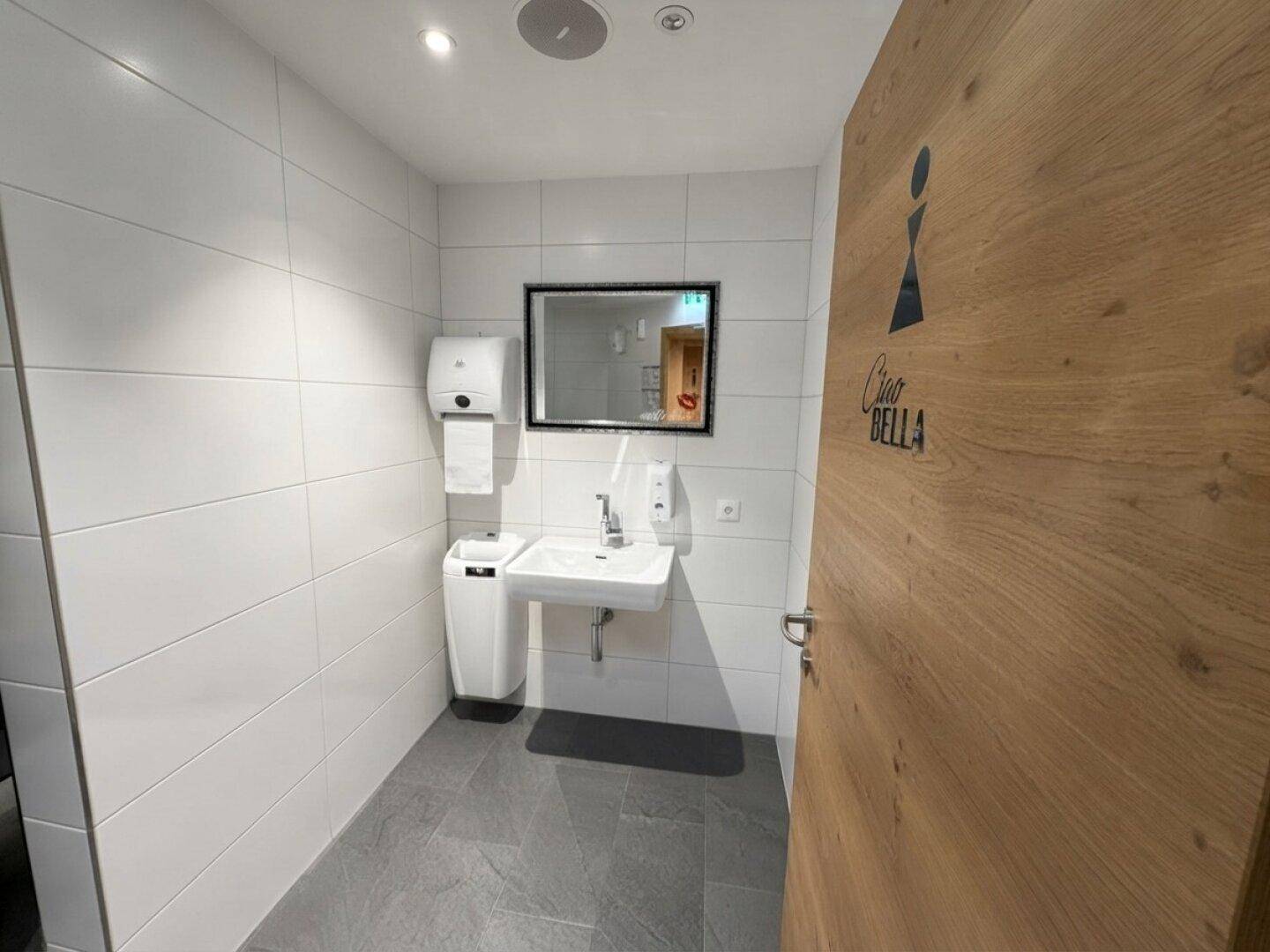Saubere und moderne Toilette mit weiß gefliesten Wänden, einem Waschbecken und einem Spiegel.