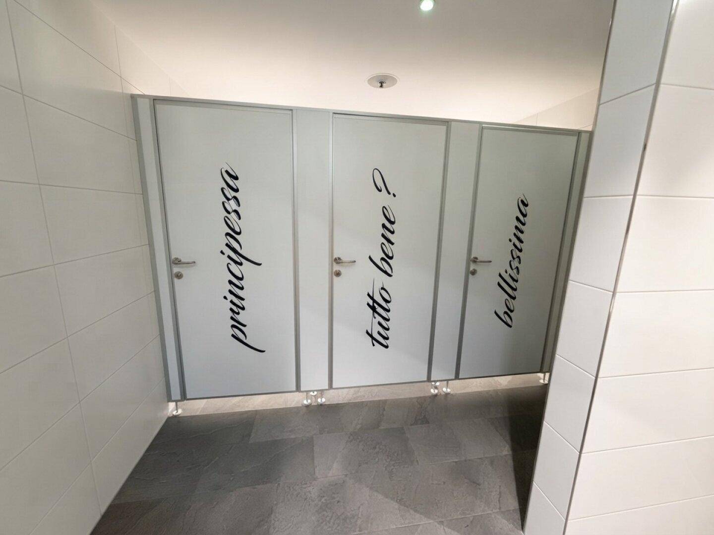 Reihe von Toilettenkabinen mit weißen Türen und dekorativen Schriftzügen in einem modernen Waschraum.
