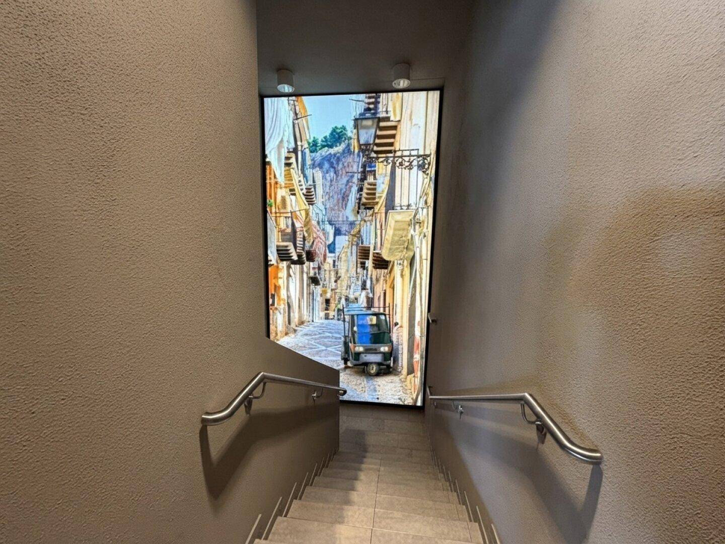 Treppenhaus mit Handläufen und einer großen, beleuchteten Wandtapete, die eine malerische Gasse darstellt.