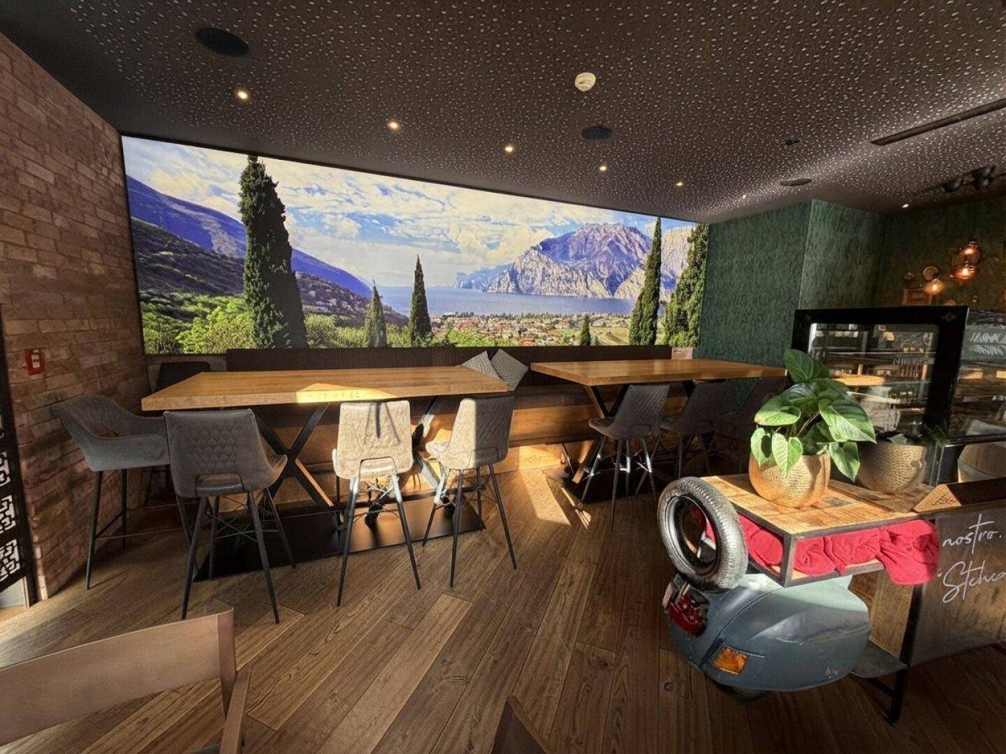 Gemütlicher Restaurantbereich mit Holztischen, gepolsterten Stühlen und einer großen Wandtapete mit Seelandschaft.