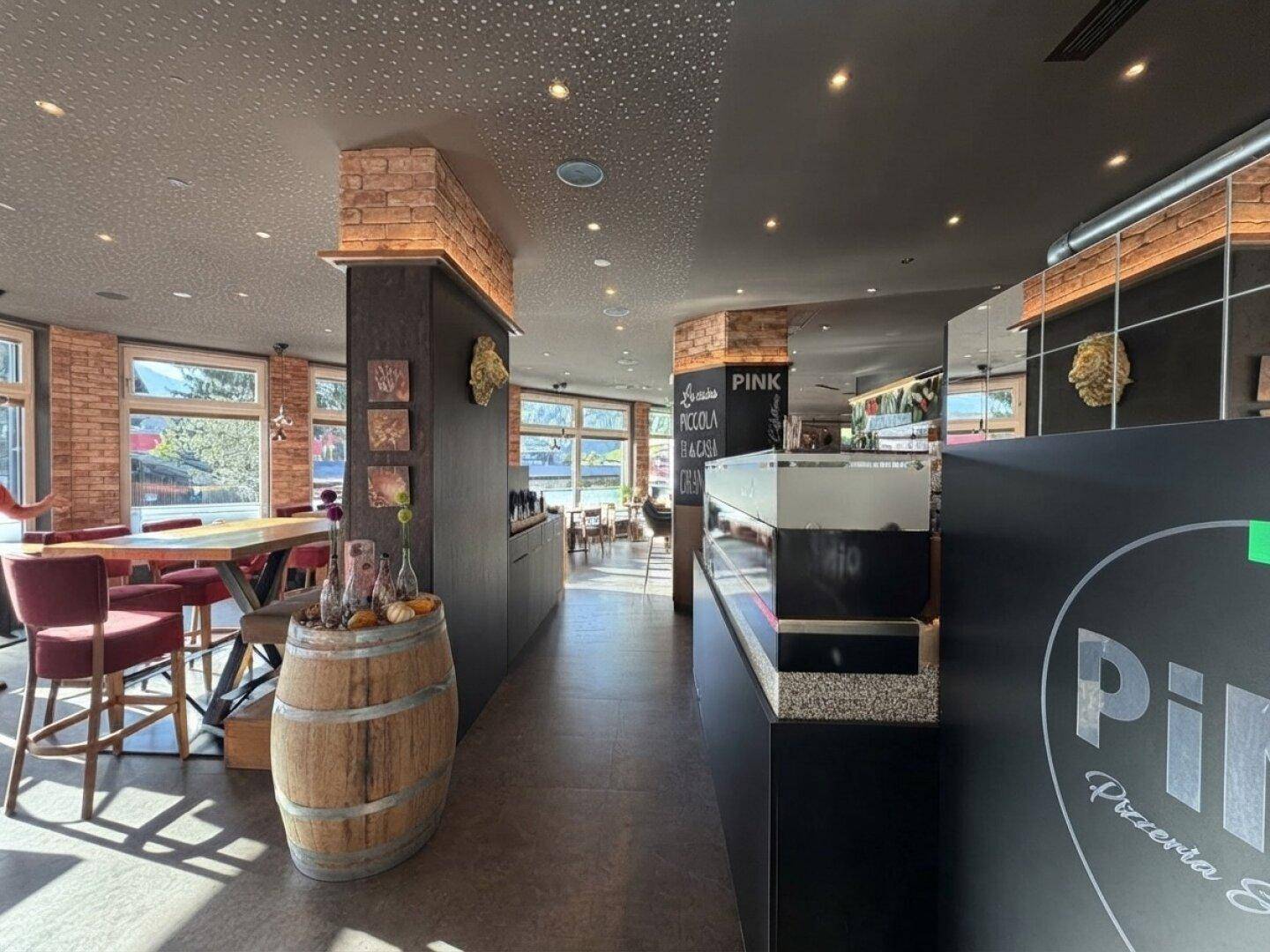 Langer Flur in einem Restaurant mit Holzboden, Ziegelpfeilern und Blick auf weitere Sitzbereiche durch große Fenster.