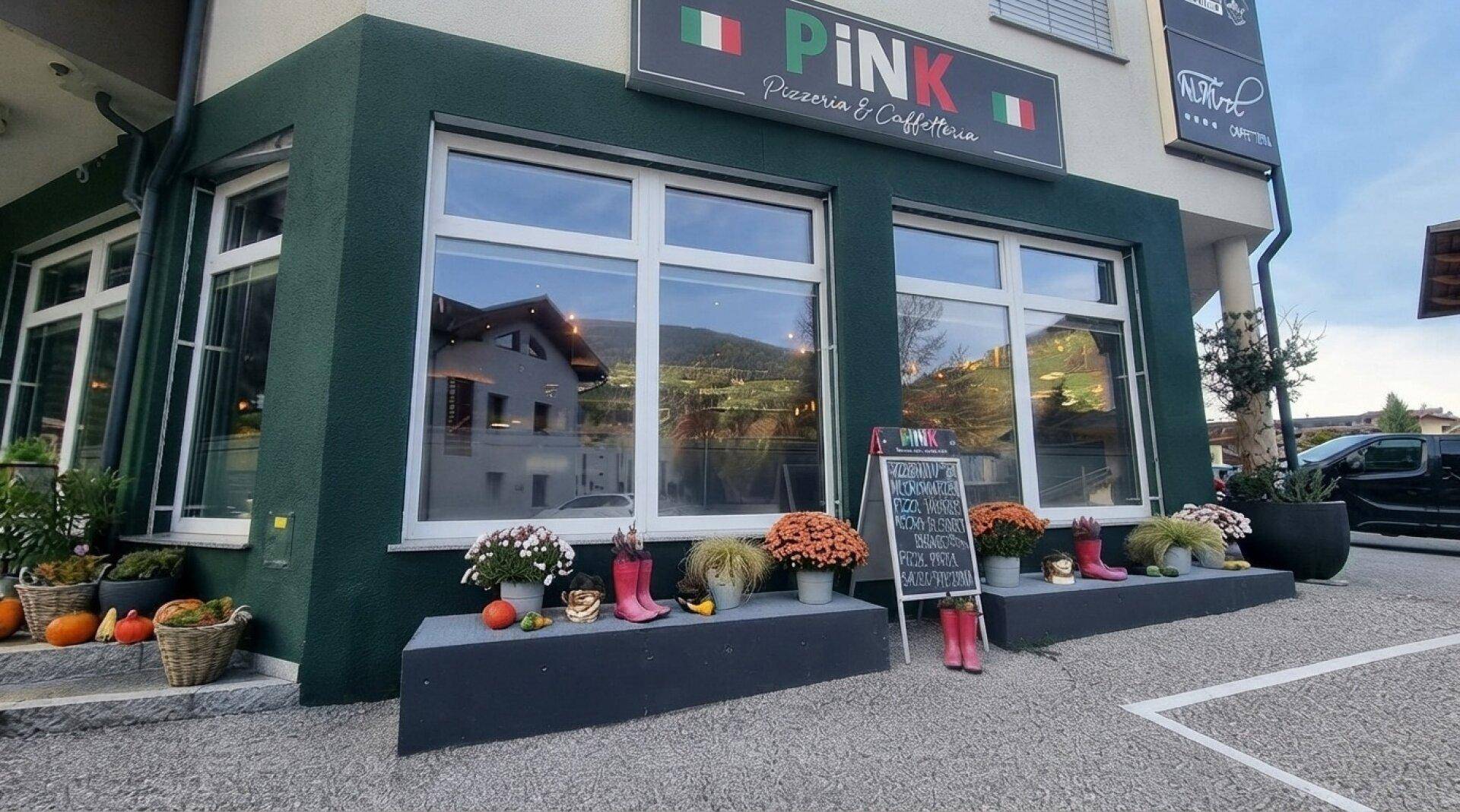 Außenansicht eines Restaurants mit grüner Fassade, großen Fenstern und saisonaler Dekoration vor dem Eingang.