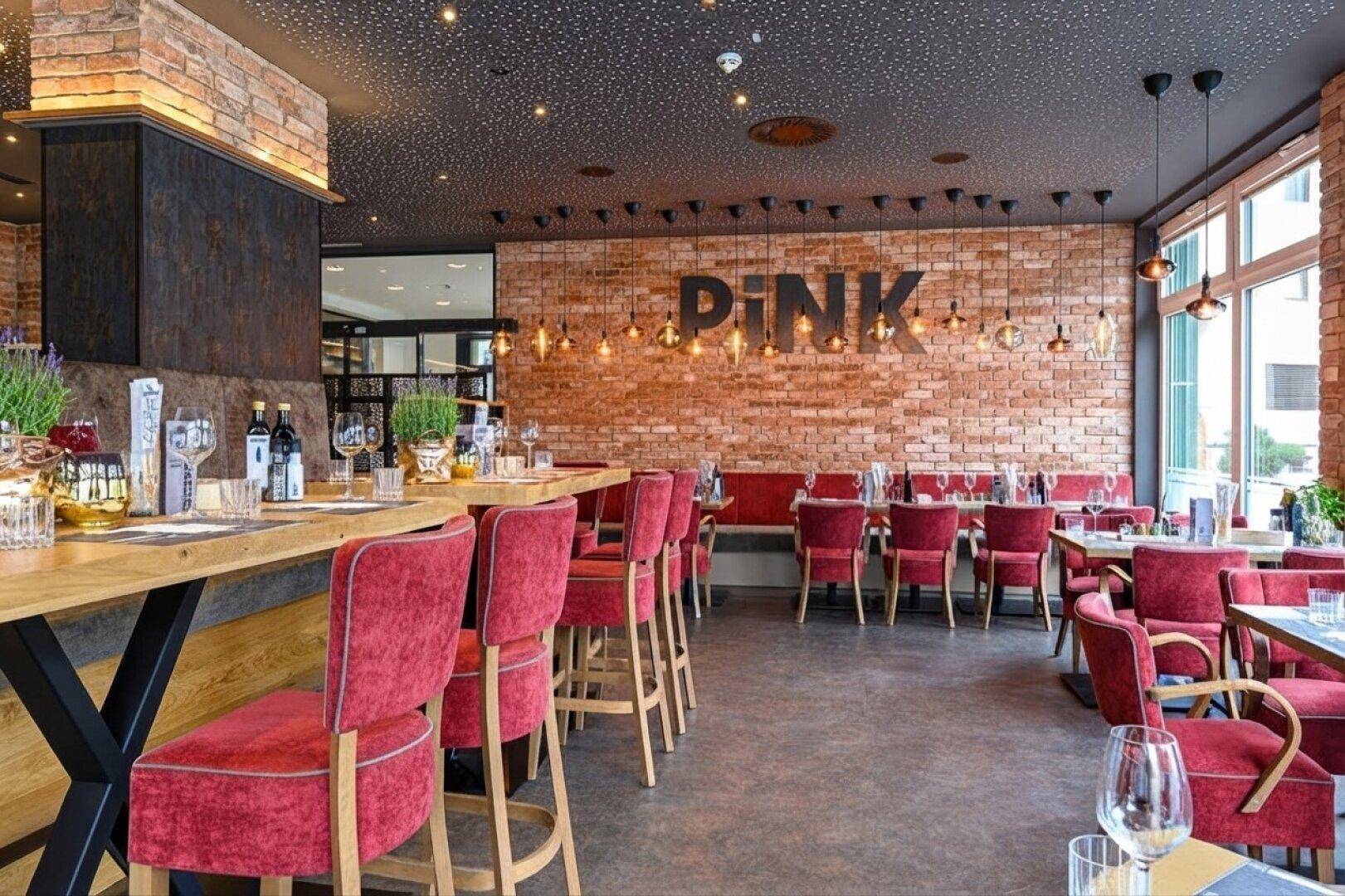 Weitläufiger Restaurantbereich mit roten Stühlen, Holztischen und einer Ziegelwand mit dem Restaurantnamen.