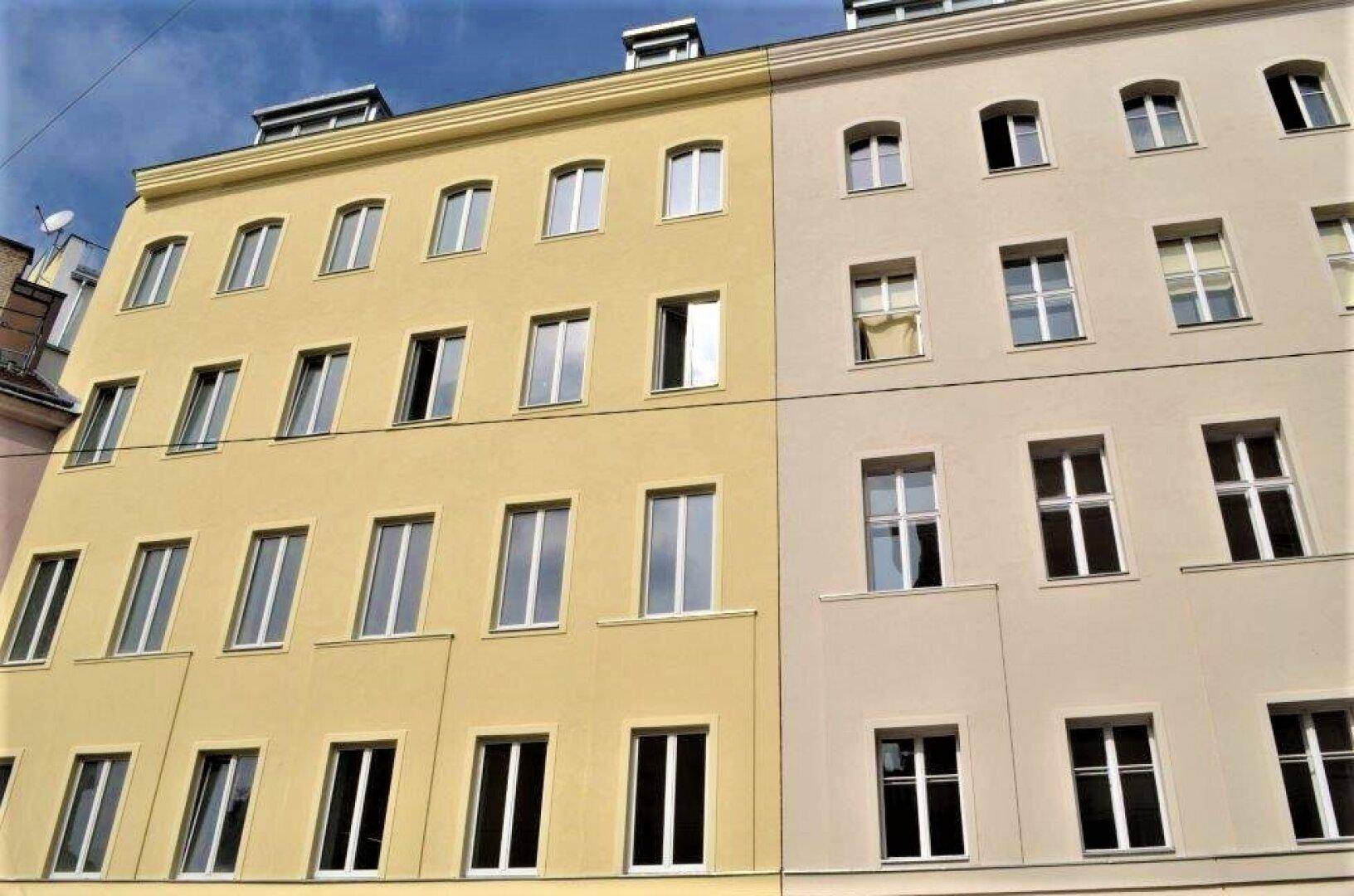 Zwei angrenzende Wohngebäude mit unterschiedlichen Fassadenfarben unter blauem Himmel in einer städtischen Umgebung.