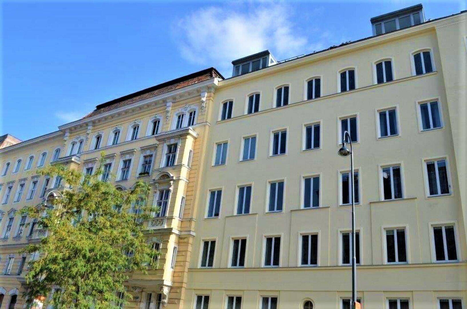 Zwei angrenzende Gebäude mit unterschiedlicher Architektur, eines davon ein Altbau mit Stuckfassade und das andere mit einer schlichten Fassade.