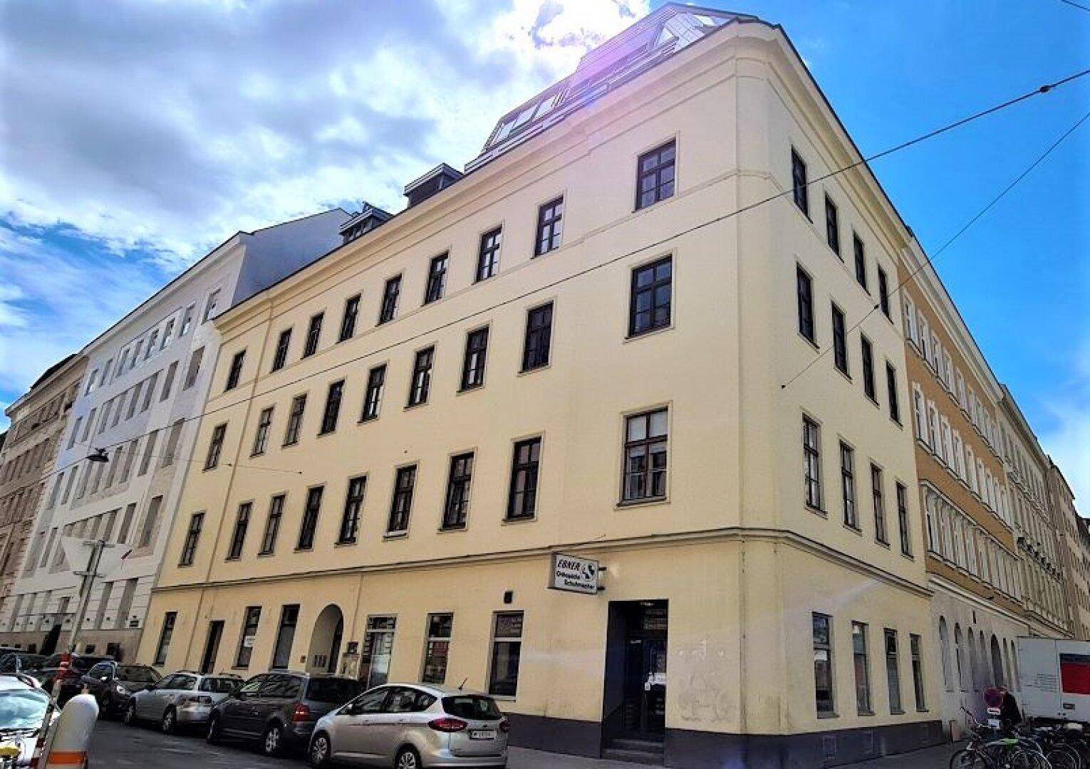 Eckgebäude mit mehreren Stockwerken und vielen Fenstern, umgeben von parkenden Autos auf der Straße.