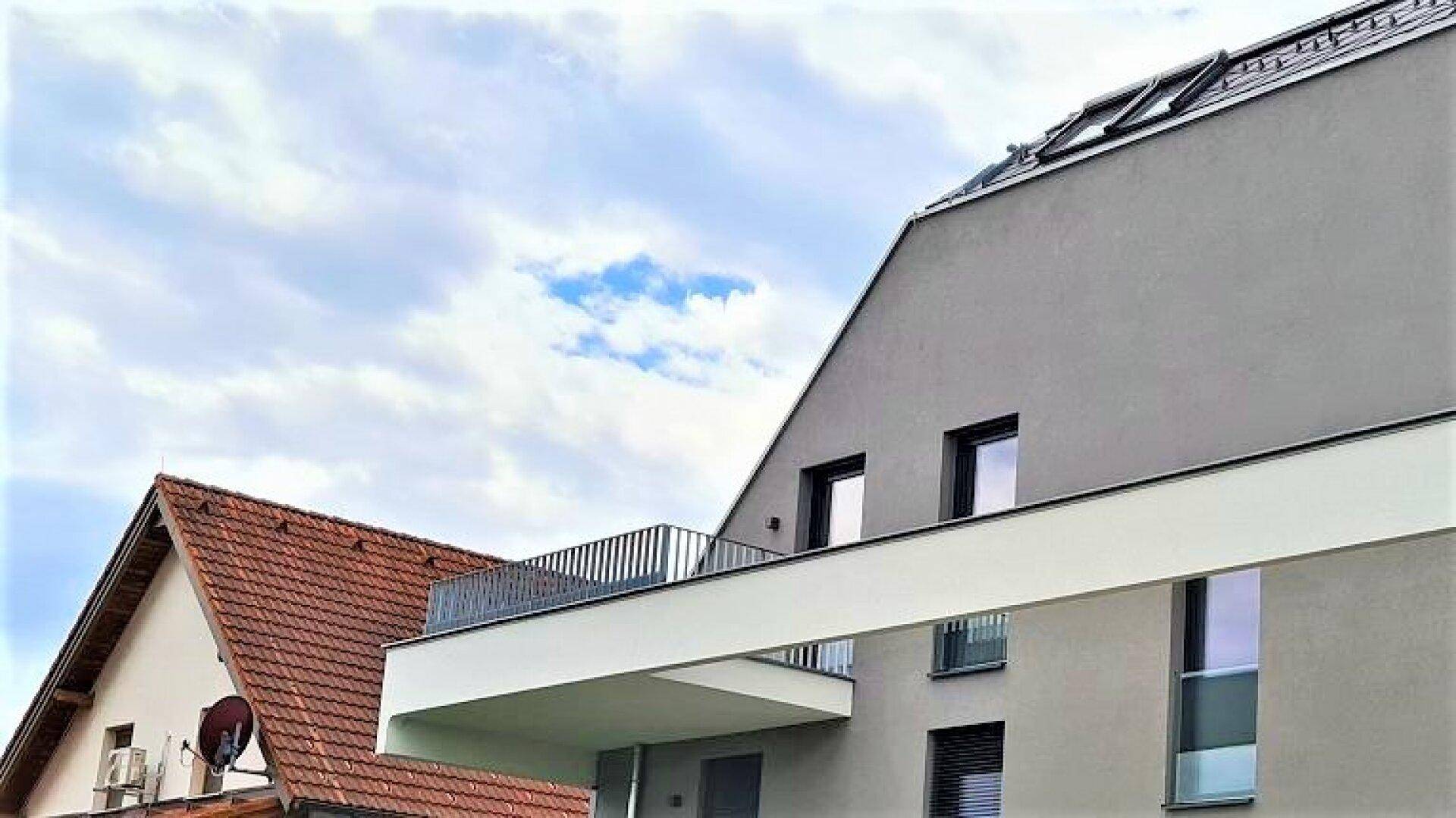 Seitenansicht eines modernen Gebäudes mit Balkon neben einem traditionellen Haus mit Ziegeldach.