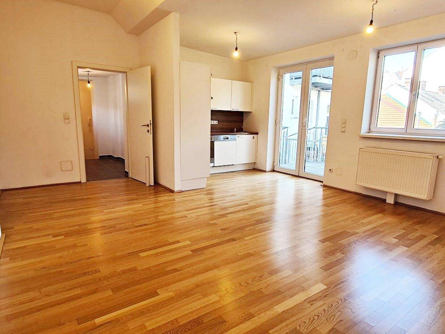 Offener Wohnbereich mit Parkettboden, Zugang zum Balkon und kleiner Küchenzeile.
