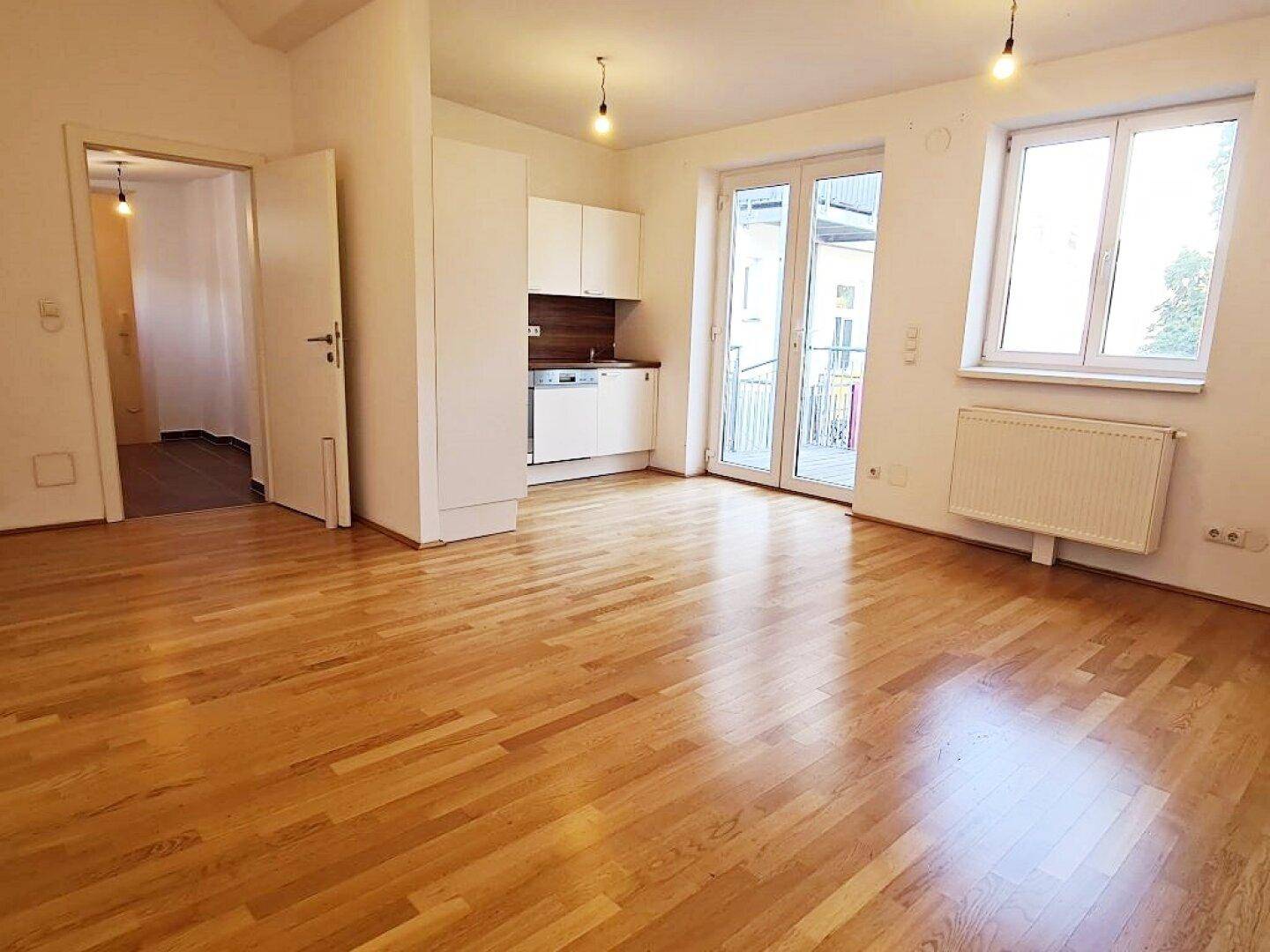 Geräumiger Wohnbereich mit Holzboden, weißer Küchenzeile und viel Tageslicht.