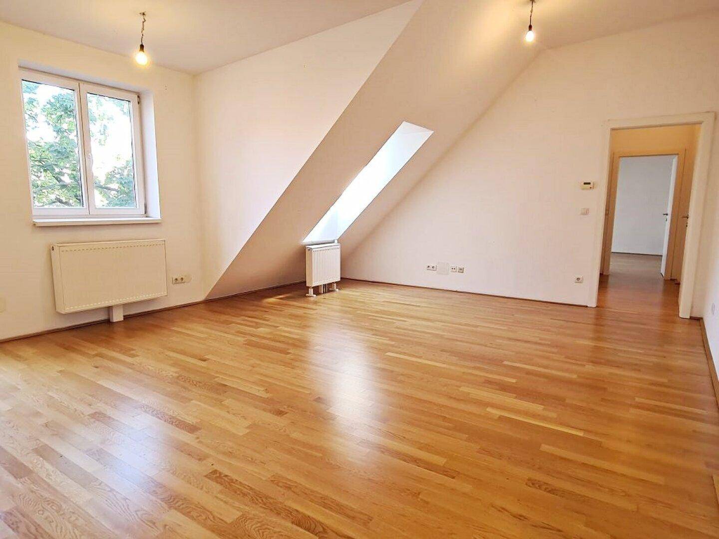 Heller Wohnraum mit Holzboden, Dachschrägen und großem Fenster mit Ausblick.