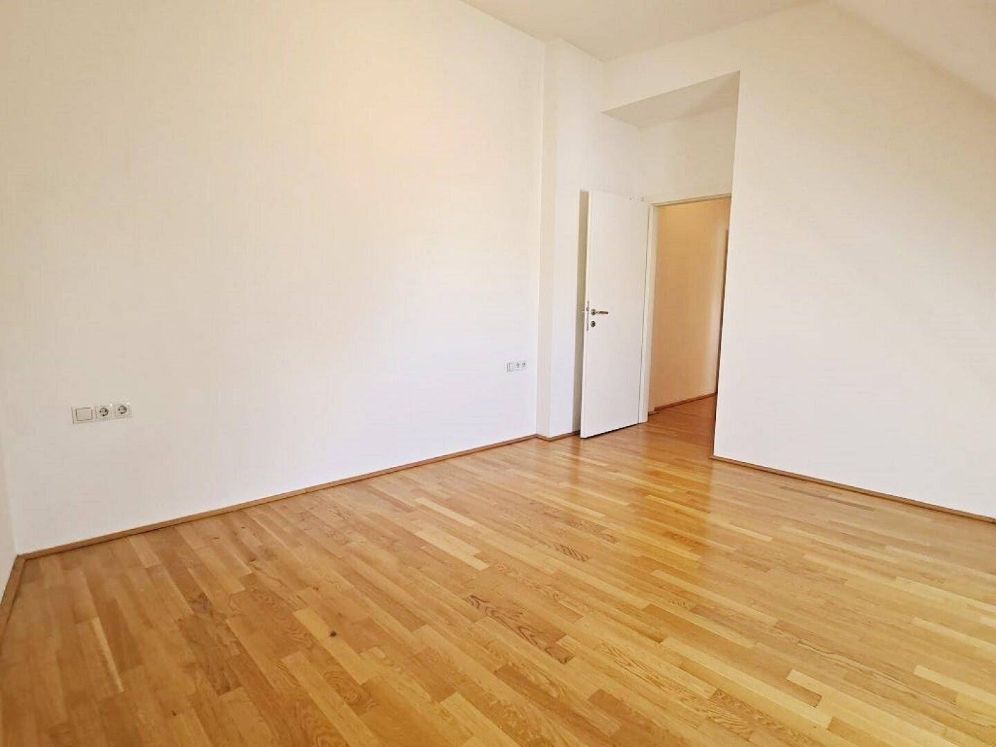 Helles Zimmer mit Holzboden und weiß gestrichenen Wänden, mit Zugang zu einem Flur.