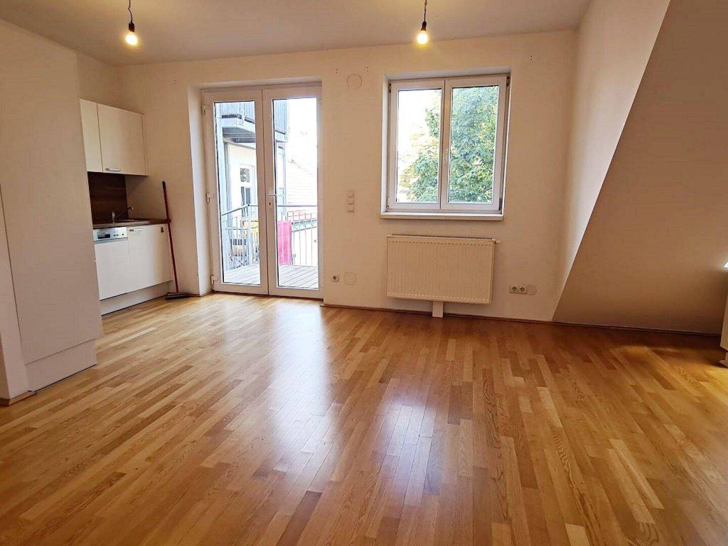 Heller Wohnbereich mit Holzboden, kleiner Küchenzeile und Zugang zum Balkon.