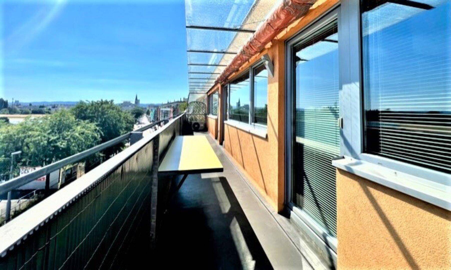 Langer Balkon mit Glasüberdachung und Blick auf die Stadt und umliegende Gebäude.