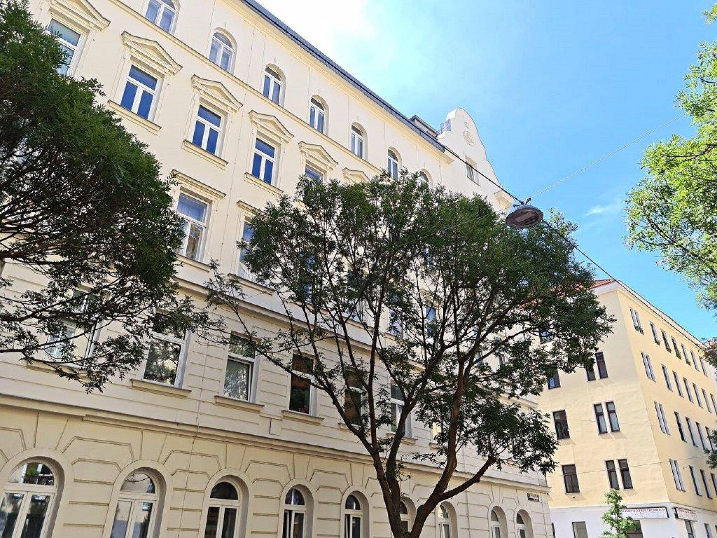 Fassade eines Gebäudes mit klassischer Architektur, teilweise von Bäumen verdeckt, unter blauem Himmel.
