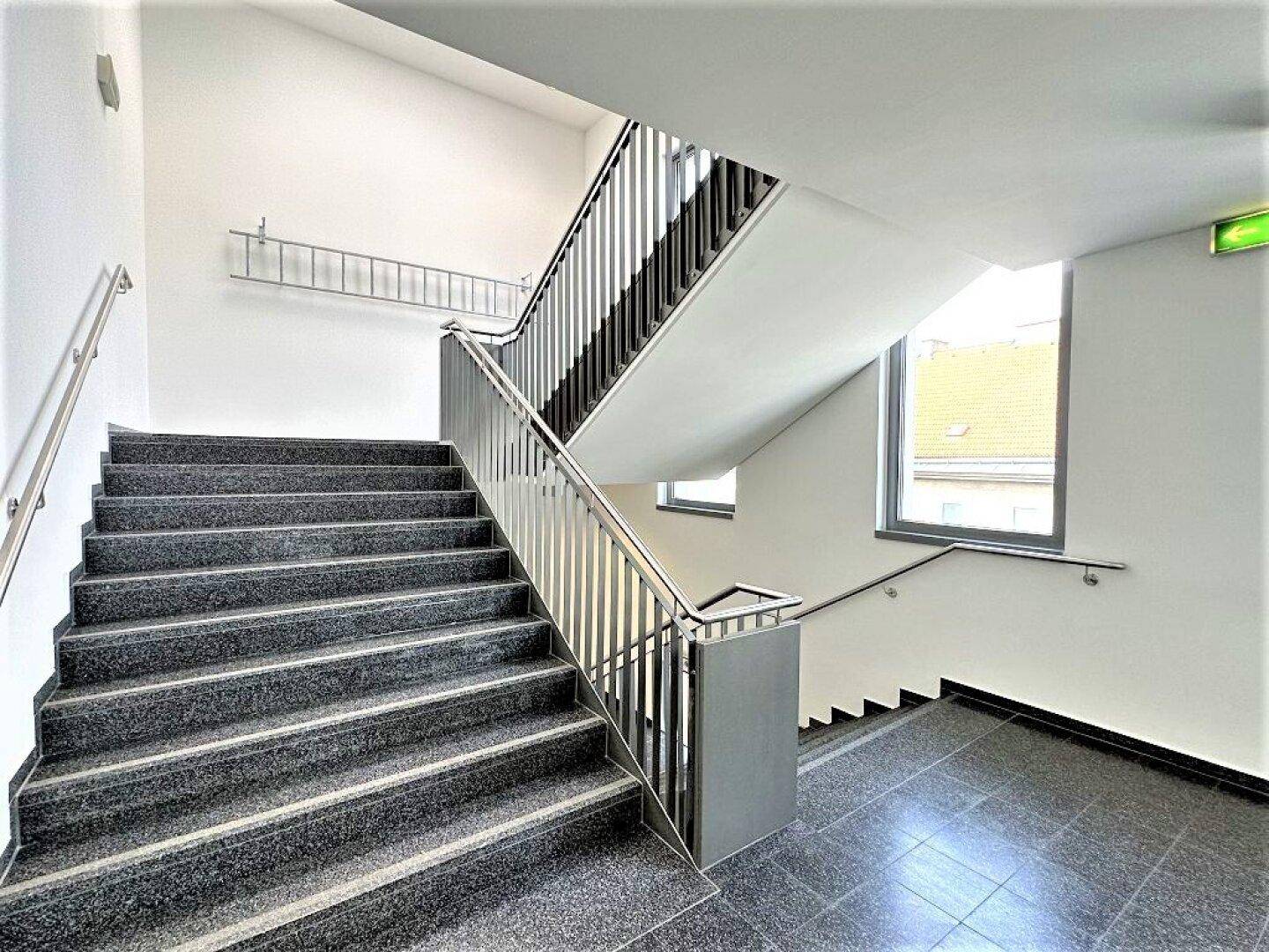 Modernes Treppenhaus mit grauen Stufen, Edelstahlgeländer und Fenstern für natürliches Licht.