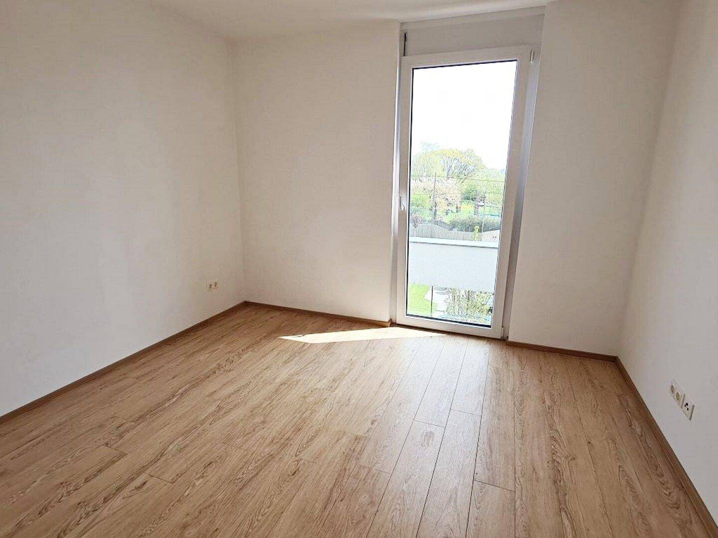 Helles Zimmer mit Holzboden und einem hohen Fenster, das viel Tageslicht hereinlässt.