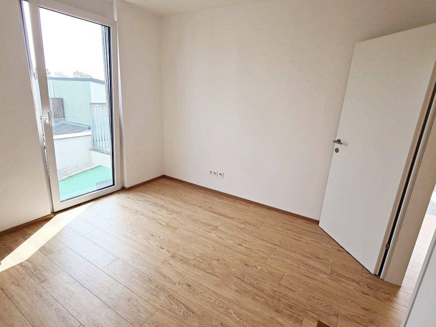 Sonniger Raum mit Holzboden und einer hohen Fenstertür, die den Raum mit Licht füllt.