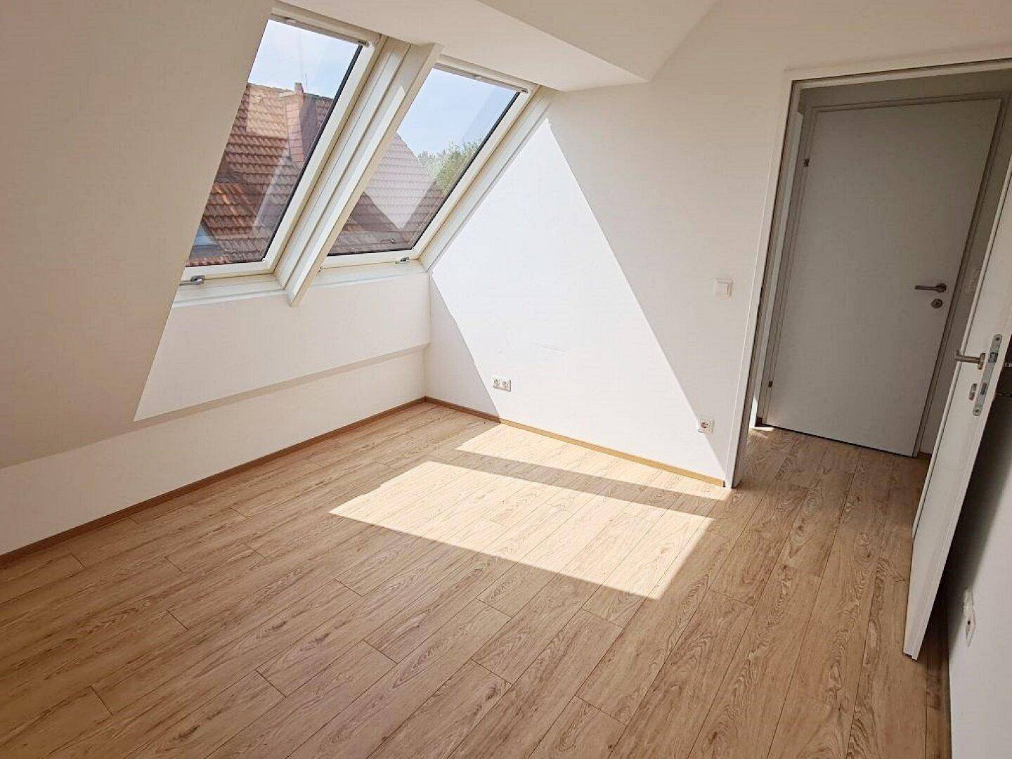 Sonniger Raum mit Holzboden, Dachschräge und zwei Dachfenstern, die viel Licht spenden.