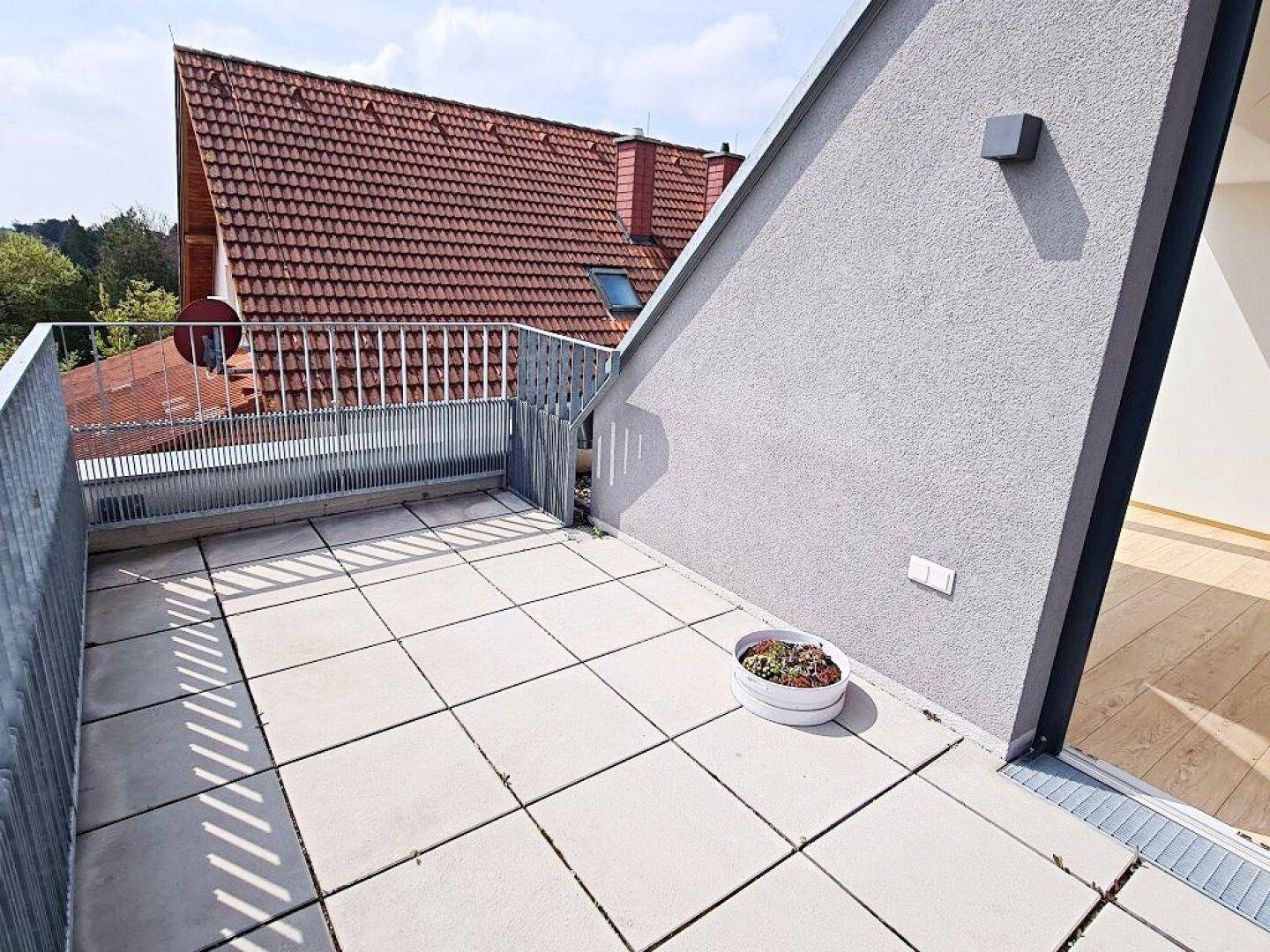 Heller Balkon mit Fliesenboden und Blick auf ein traditionelles rotes Ziegeldach in der Ferne.