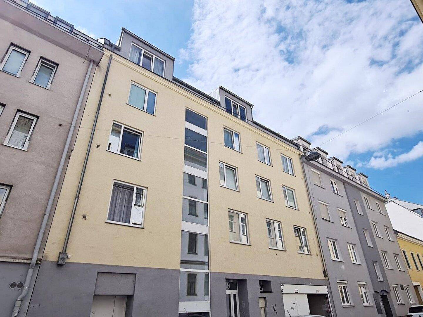 Gebäudefassade eines Wohnhauses mit beige gestrichener Außenwand und angrenzenden Gebäuden unter blauem Himmel.