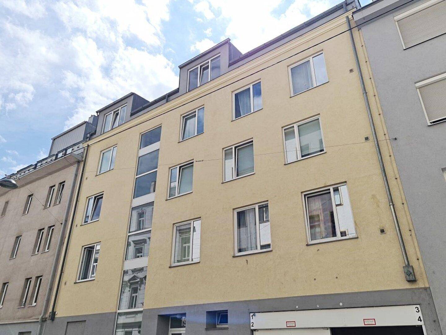 Fassade eines Mehrfamilienhauses mit hellgelbem Putz, modernen Fenstern und integriertem Aufzugsschacht.