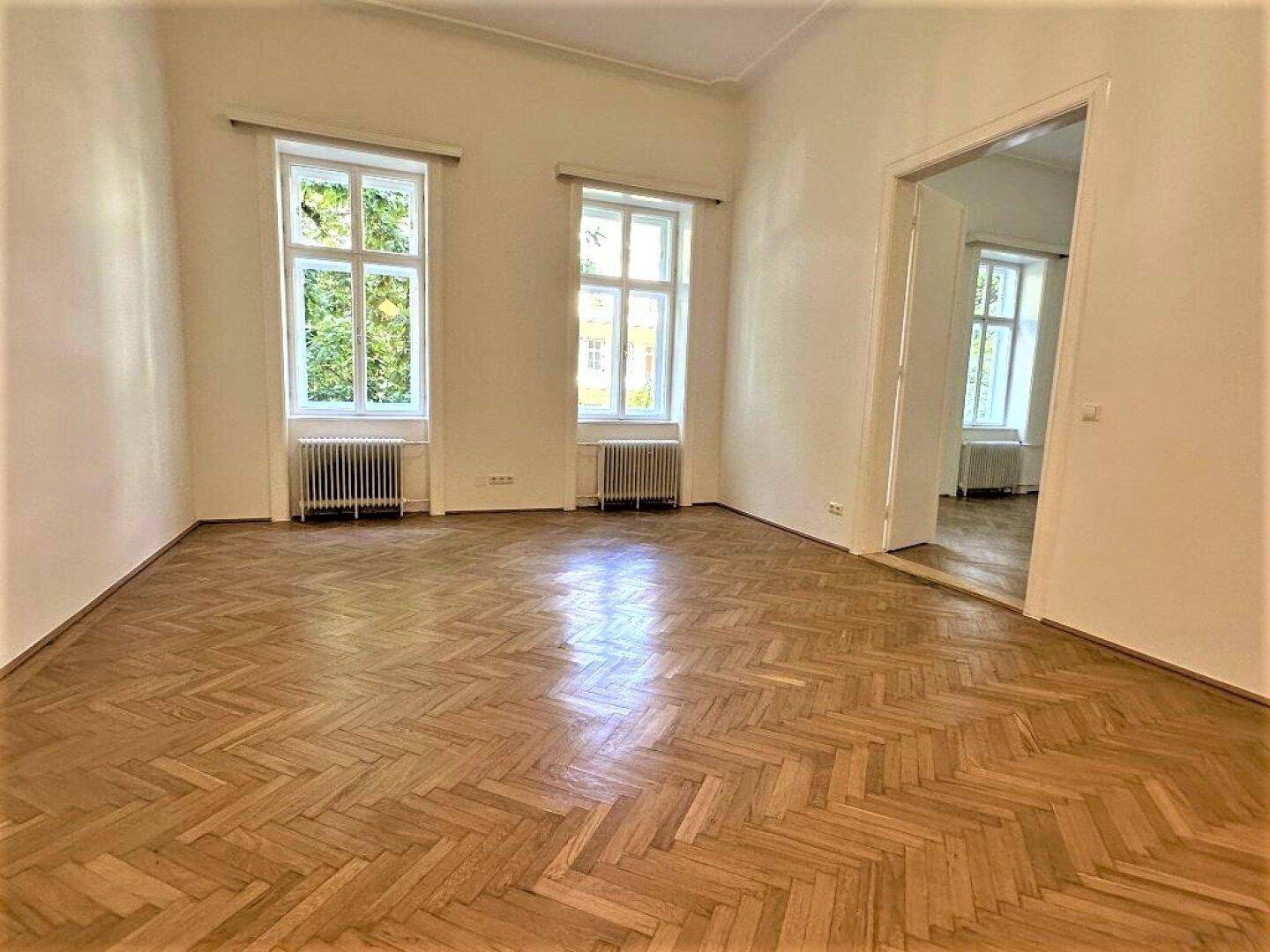 Geräumiger Wohnbereich mit Fischgrätparkett, hohen Decken und zwei großen Fenstern.