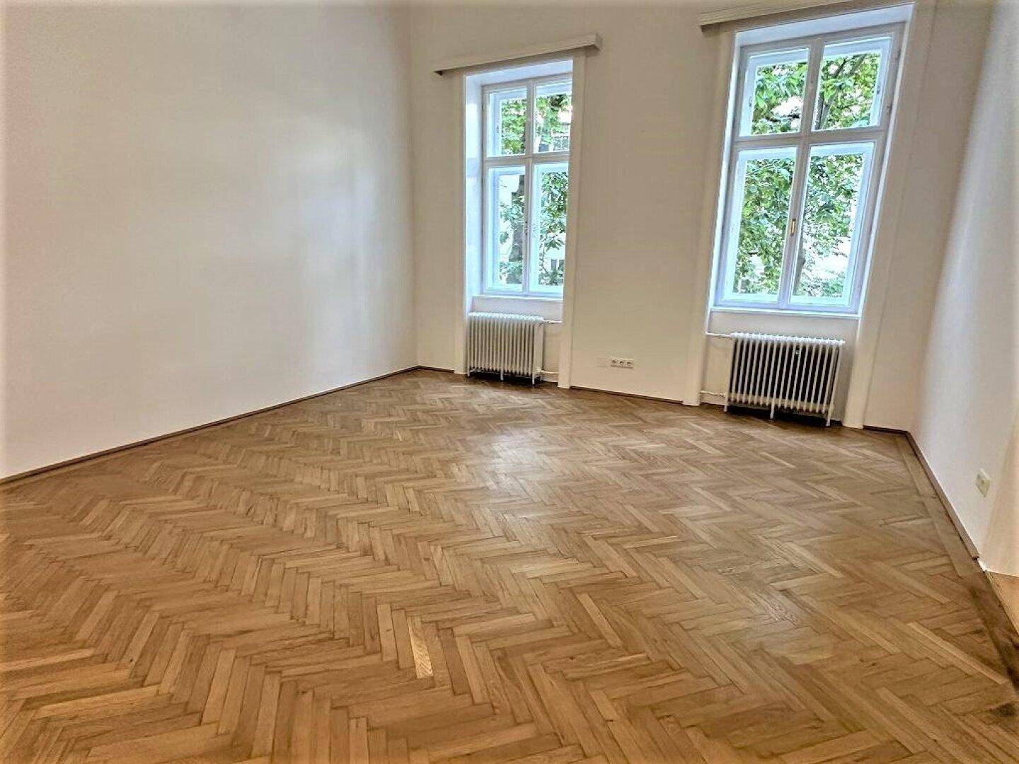 Heller Wohnraum mit Fischgrätparkett, hohen Decken und zwei großen Fenstern.