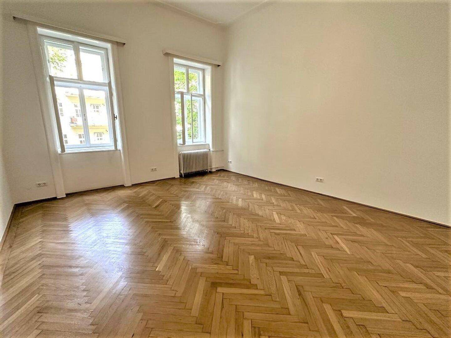 Geräumiger Raum mit Fischgrätparkett und zwei großen Fenstern, die viel Tageslicht hereinlassen.