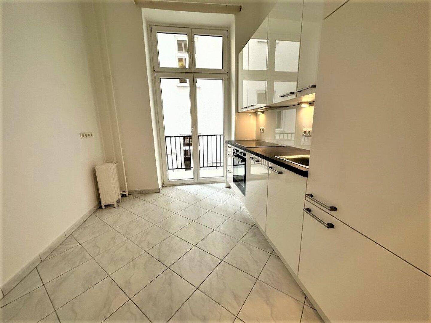 Moderne Küche mit weißen Hochglanzschränken, Fliesenboden und Zugang zum Balkon.