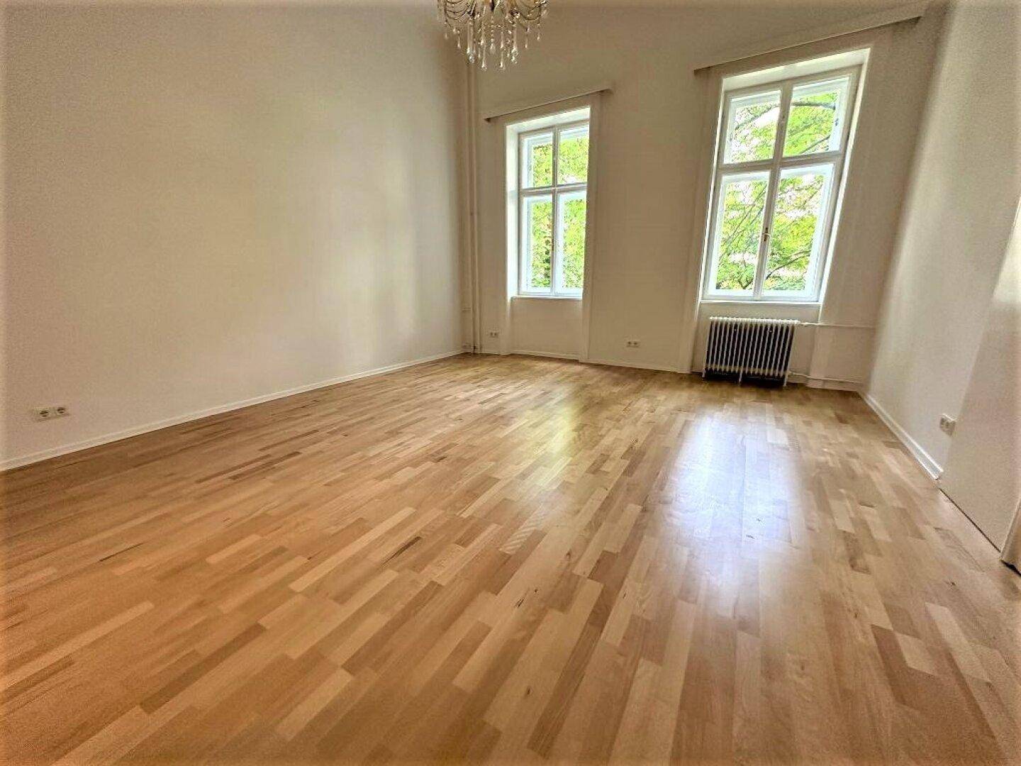 Heller Wohnraum mit hellem Parkettboden, Kronleuchter und zwei großen Fenstern.