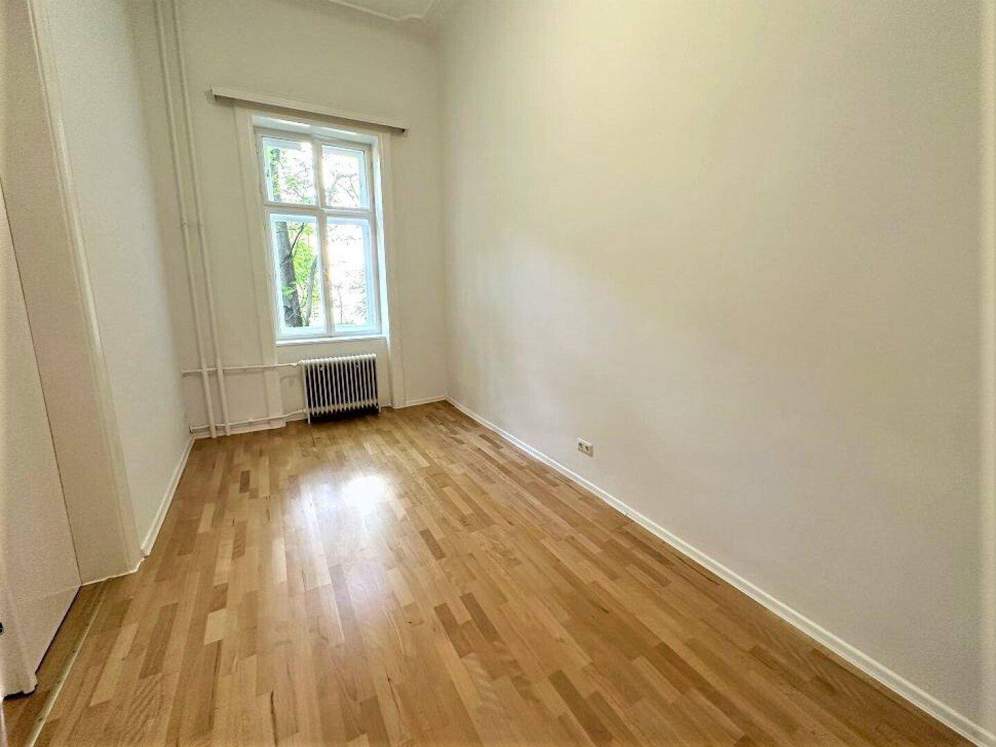 Helles Schlafzimmer mit hellem Parkettboden und einem großen Fenster mit Blick ins Grüne.